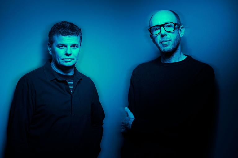 English electronic music duo The Chemical Brothers posing together against a dark blue background with atmospheric lighting
